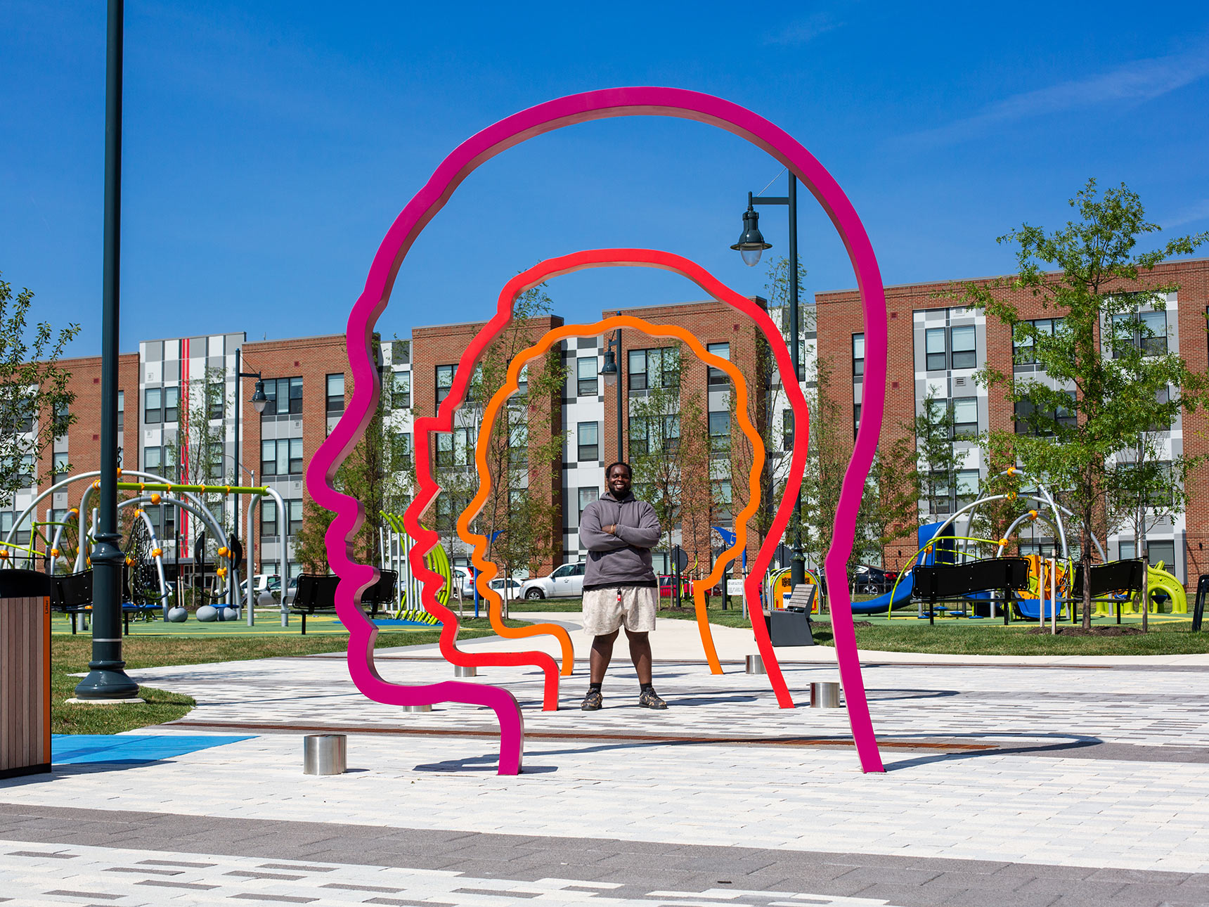 A man stands smiling in the middle of Harmonic Time public art installation is a new contemporary monument composed of a colonnade of three colorful monumental 12-foot-tall silhouette portraits in a gradient of orange, red, and purple.