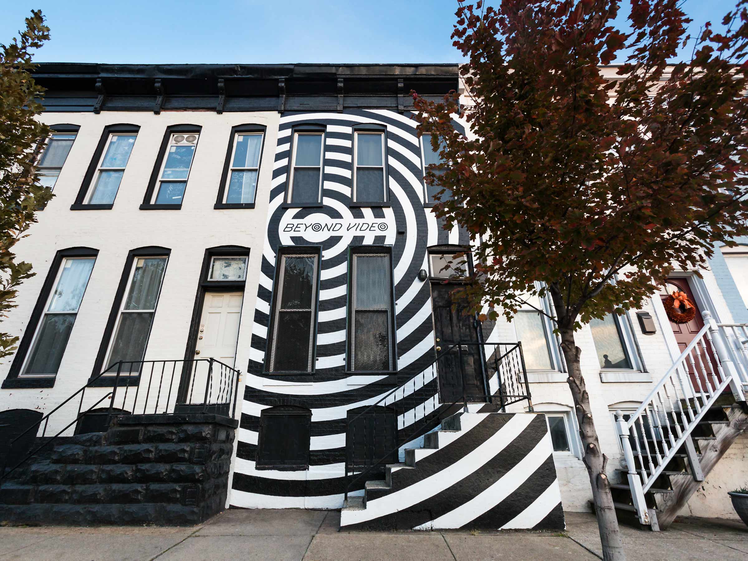 A high-contrast supergraphic black and white mural covering the entire facade of a Baltimore rowhouse with giant black concentric rings, the storefront for Beyond Video, a contemporary video store and lending library