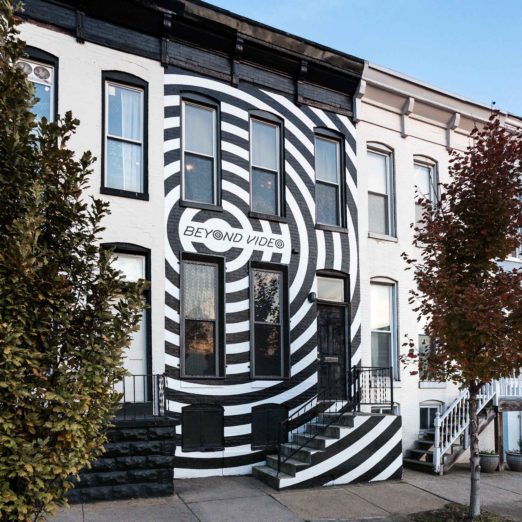 A high-contrast supergraphic black and white mural covering the entire facade of a Baltimore rowhouse with giant black concentric rings, the storefront for Beyond Video, a contemporary video store and lending library
