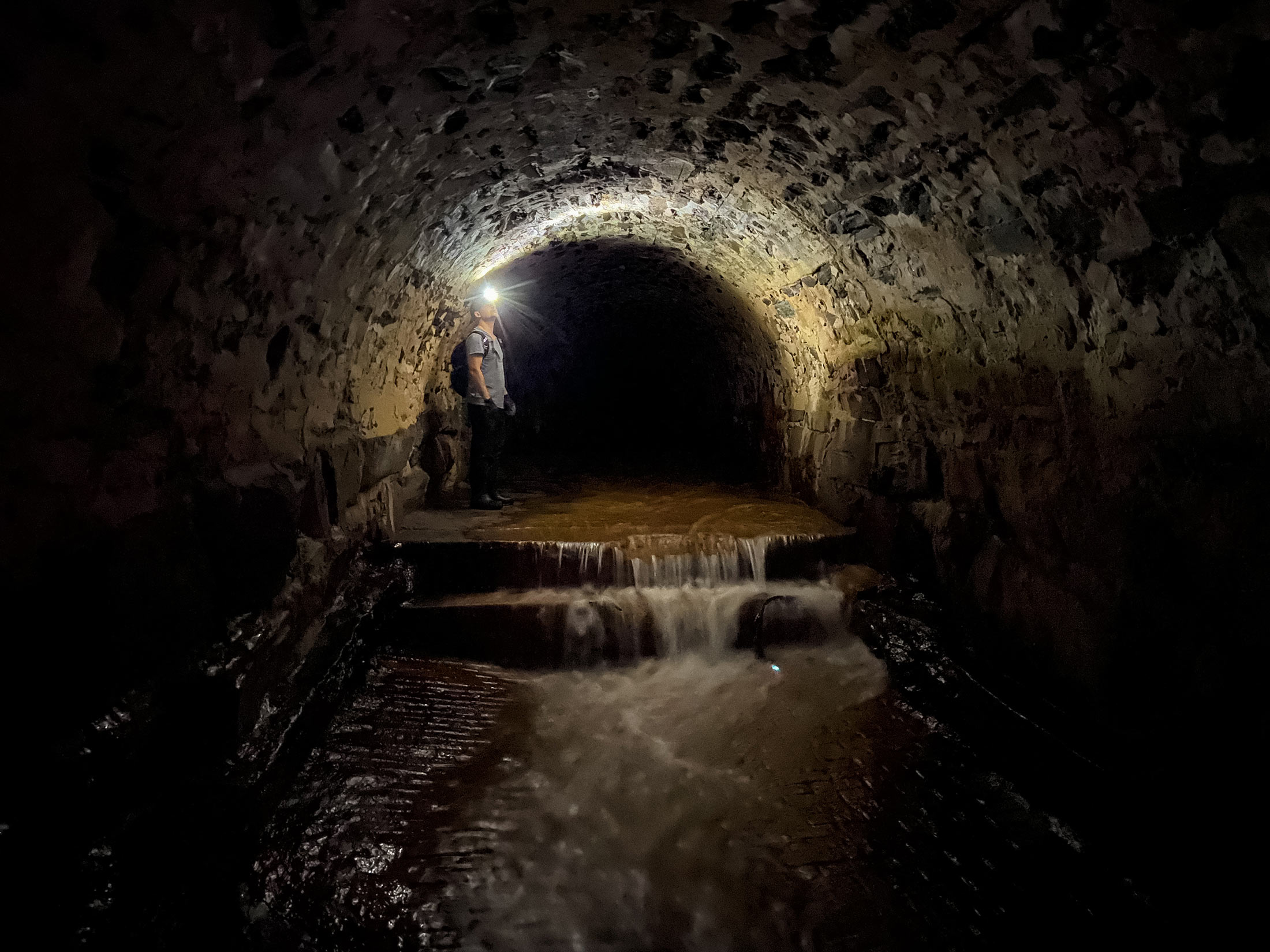 Photos of a man with a headlamp and waders standing in an underground tunnel with an arched stone ceiling. Water cascades through the tunnel down a set of steps.