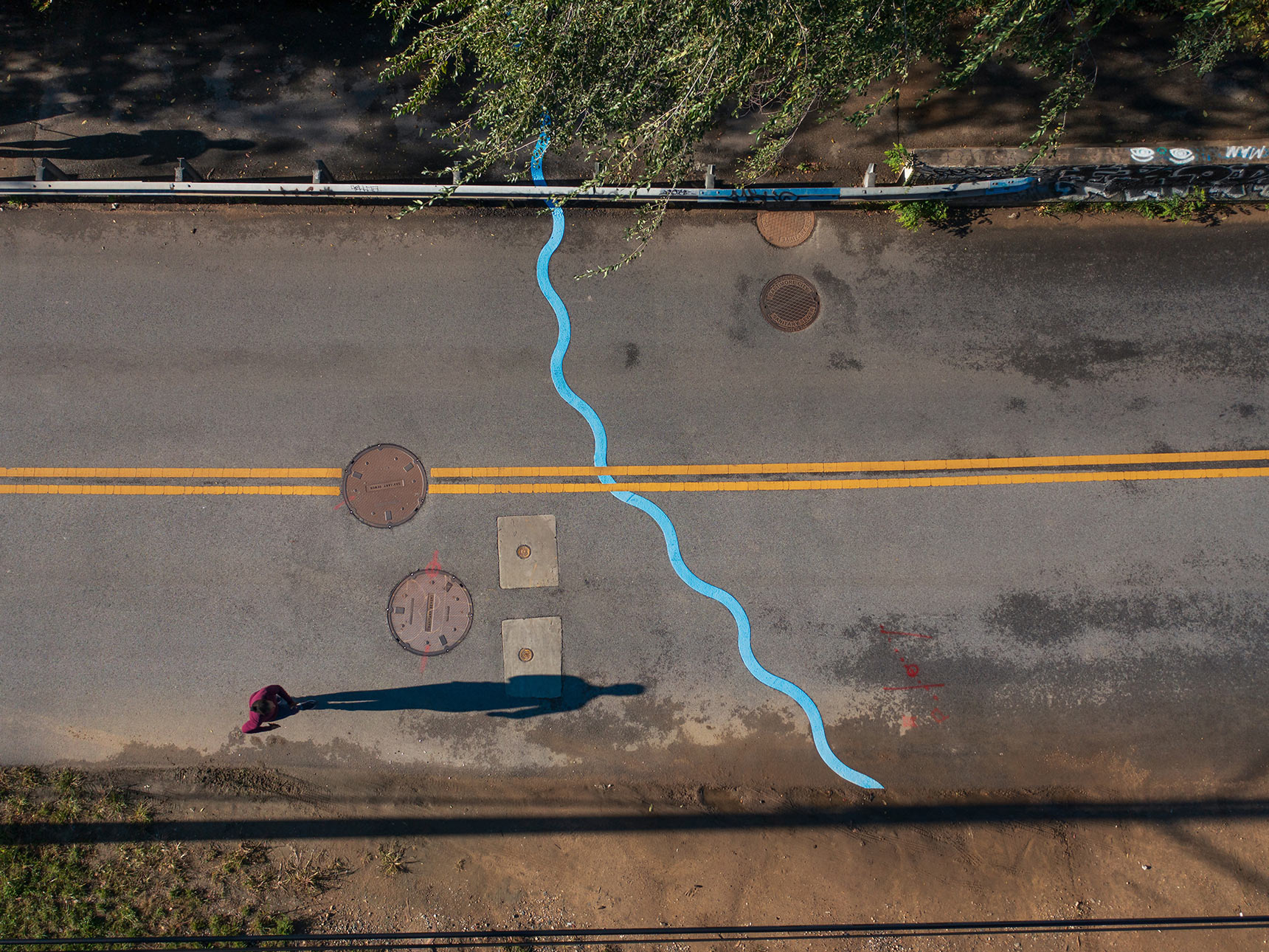 Aerial photo of a public art installation mapping the path of an underground stream. A bright blue wavy line snakes all the way across a roadway and sidewalks. The blue line appears to duck below yellow road markings. A jogger passes by casting a long early-morning shadow.