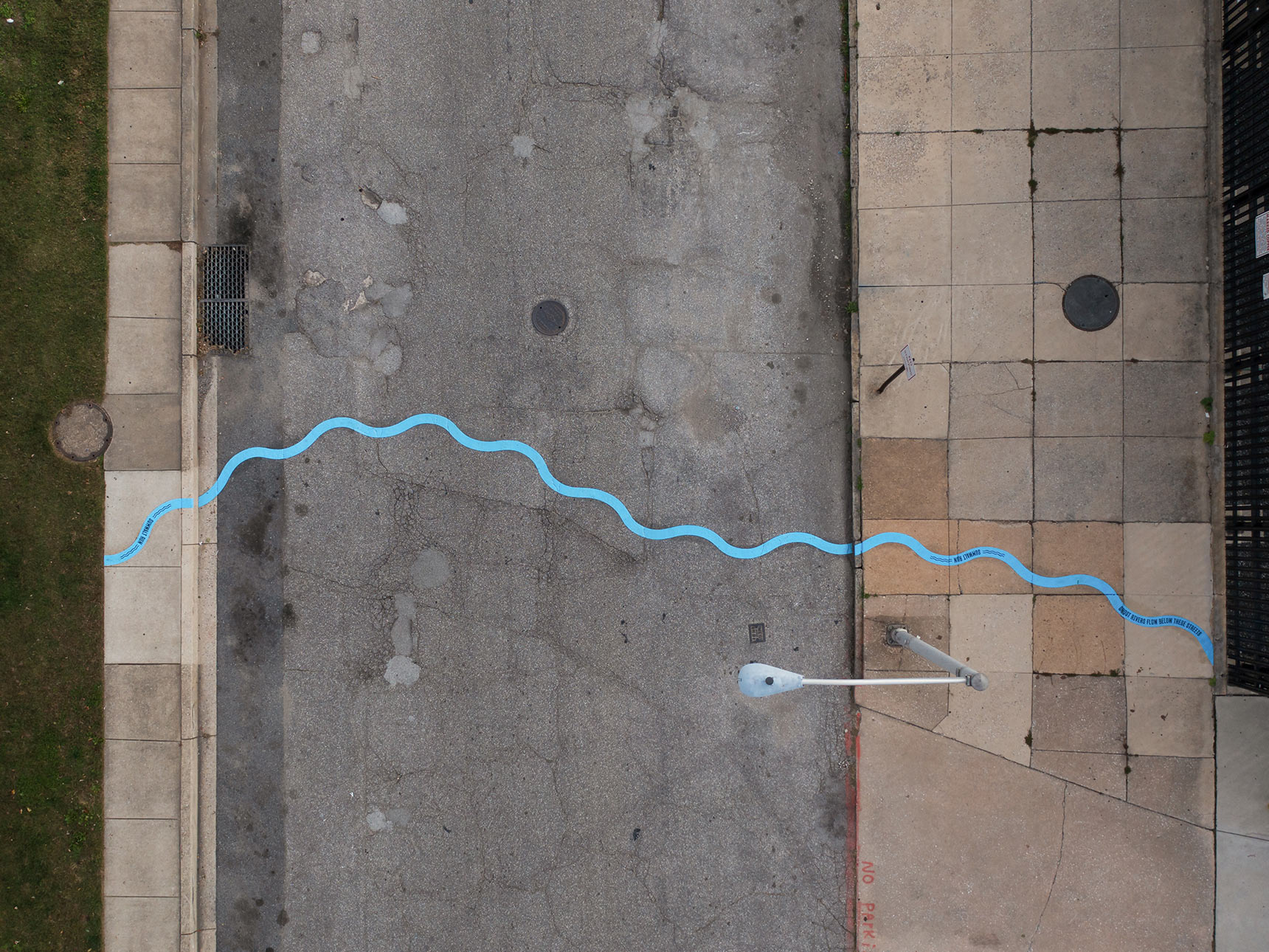 Aerial photo of a public art installation mapping the path of an underground stream. A bright blue wavy line snakes all the way across a roadway and wide sidewalk.