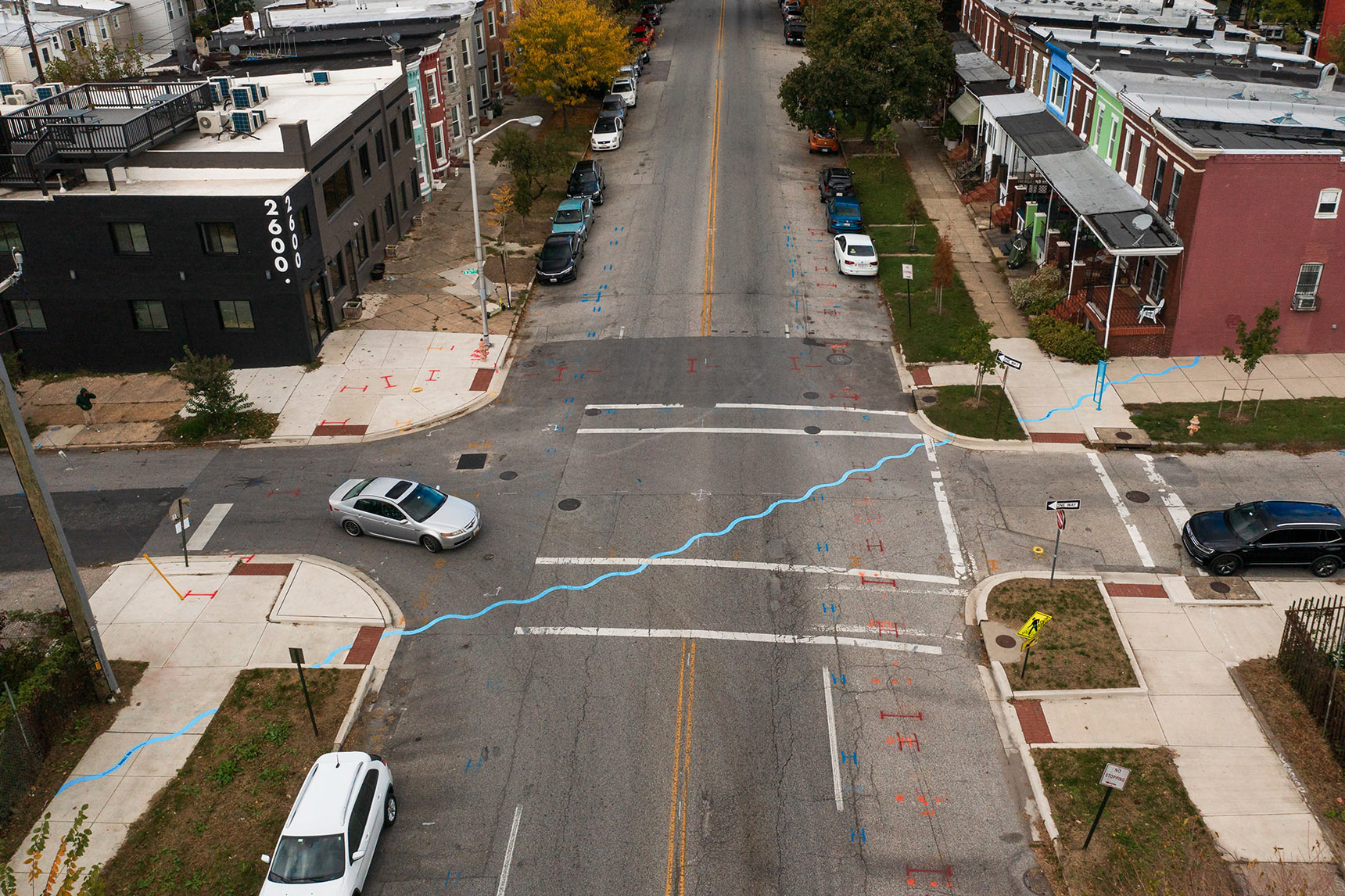 Aerial photo of a public art installation mapping the path of an underground stream. A bright blue wavy line snakes diagonally across an intersection and sidewalks, dead-ending into the side of a block of Baltimore rowhouses.