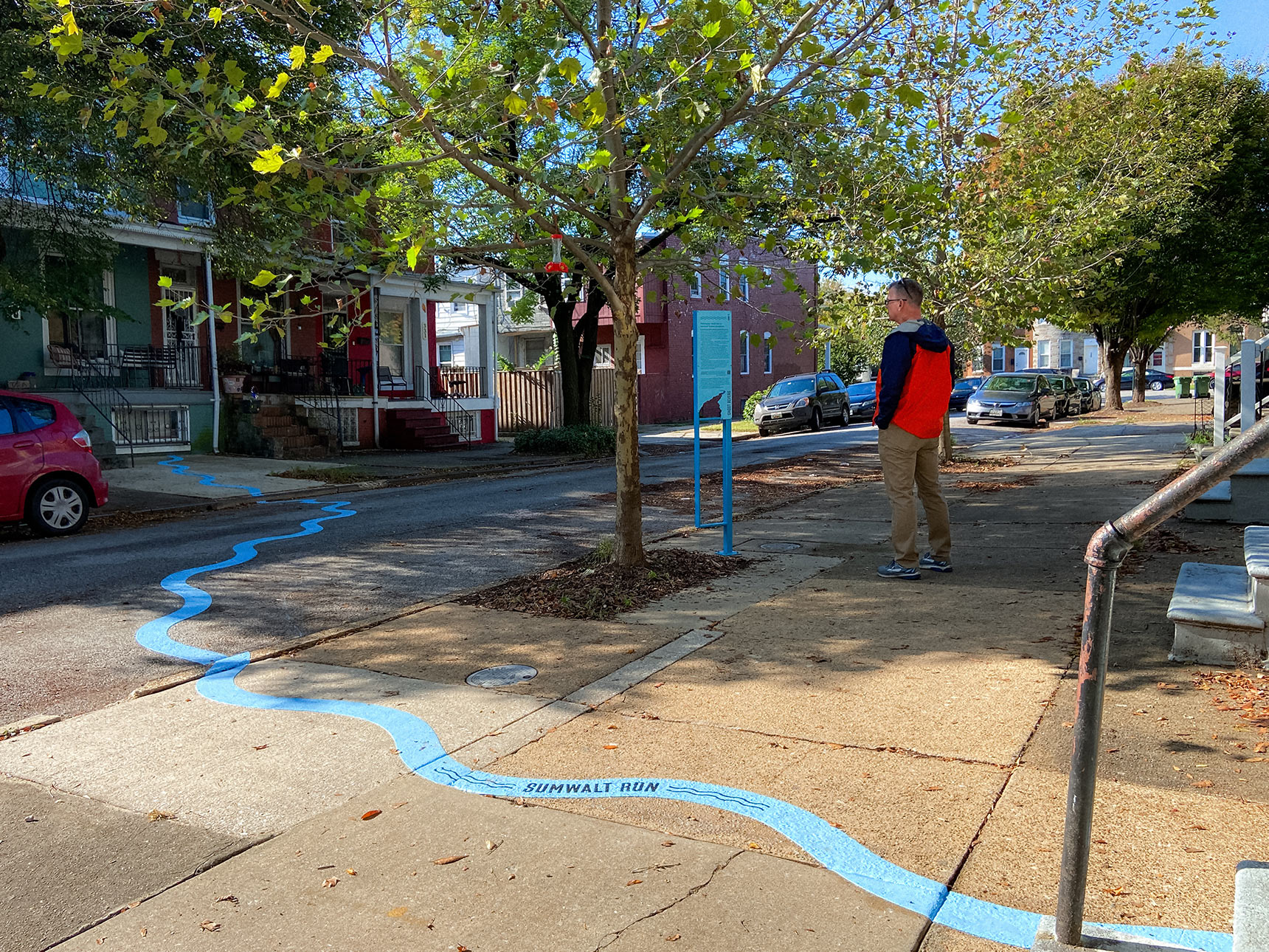 A man stands on a sidewalk in a residential neighborhood, reading a blue interpretive sign for a public art installation mapping a buried stream in Baltimore. Photo shows a wide sidewalk and street, with a six-inch wide bright blue wavy line snaking across. The words Sumwalt Run are imprinted onto the line.