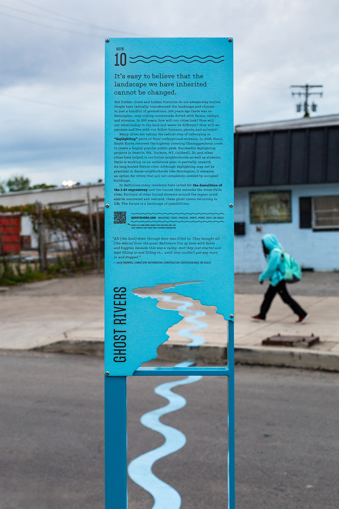 Photo of a public art installation mapping the path of an underground stream onto the pavement, showing a tall blue interpretive sign, with the form of a river cut out from the bottom of the sign panel. Through the gap in the sign, a bright blue wavy line snakes across the street.