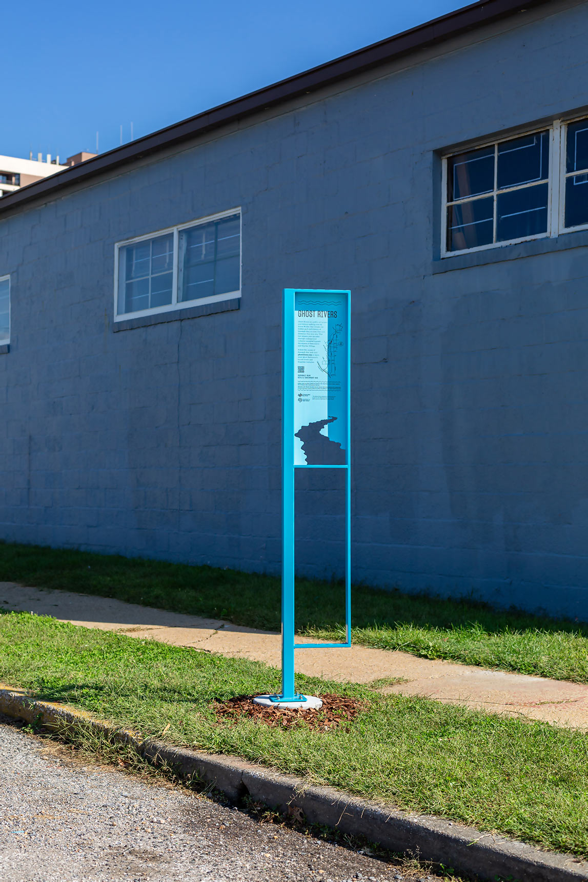 Photo of a public art installation mapping the path of an underground stream onto the pavement, showing a tall blue interpretive sign, standing in a grass strip next to a sidewalk with the form of a river cut out from the bottom of the sign panel.