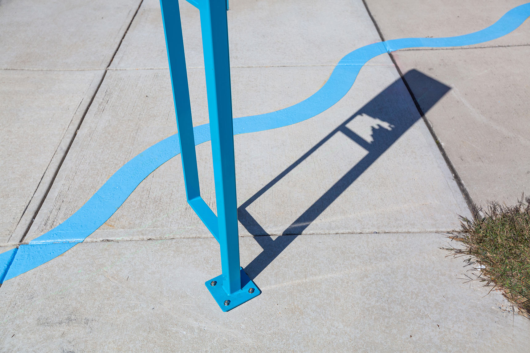 Detail photo of a public art installation mapping the path of an underground stream. A bright blue wavy line snakes across a sidewalk behind the distinctive shadow of a sculptural interpretive sign.