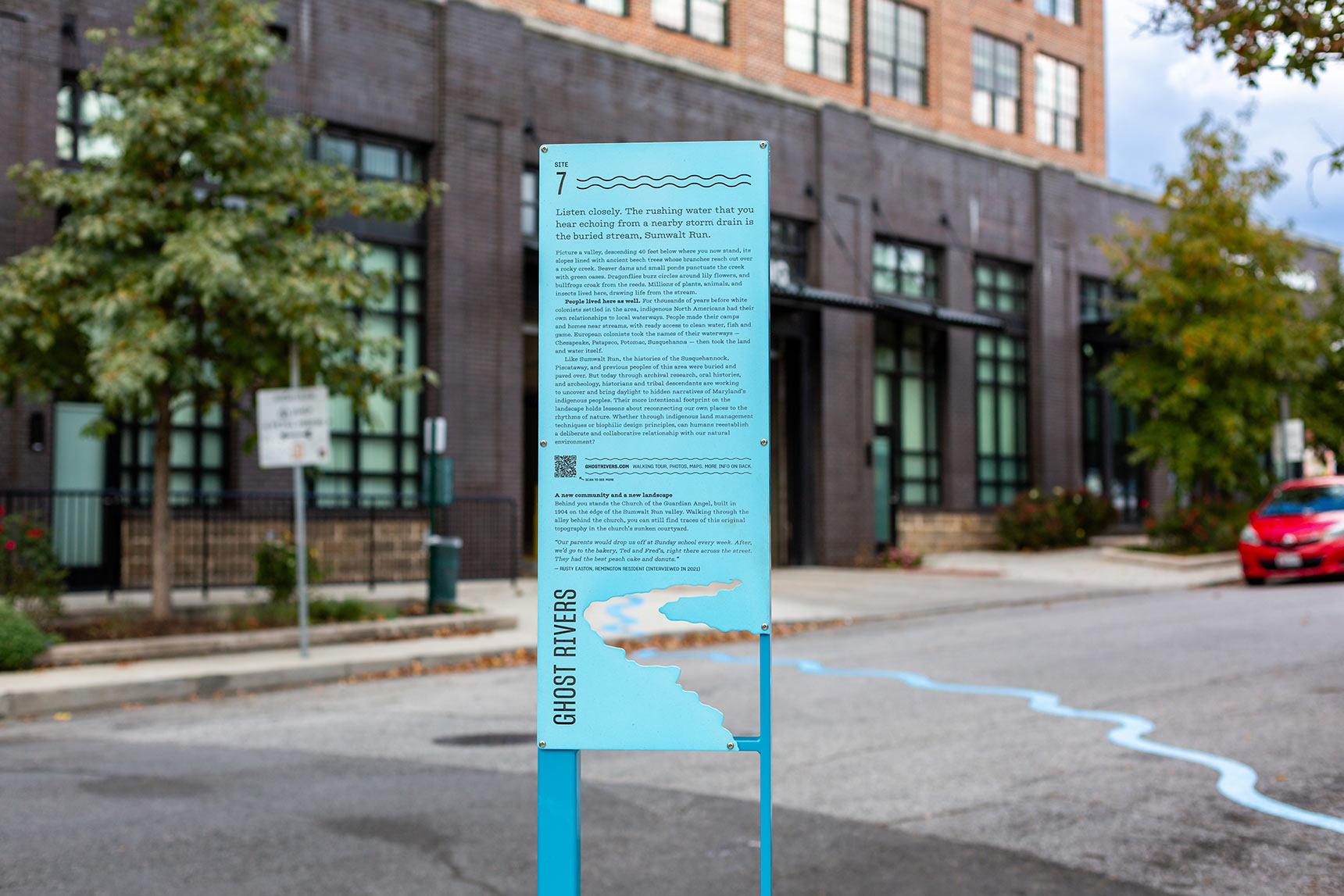 A photo of a tall blue interpretive sign, with the form of a river cut out from the bottom of the sign panel. Through the gap in the sign, a bright blue wavy line snakes across the street, with an apartment building in the background.