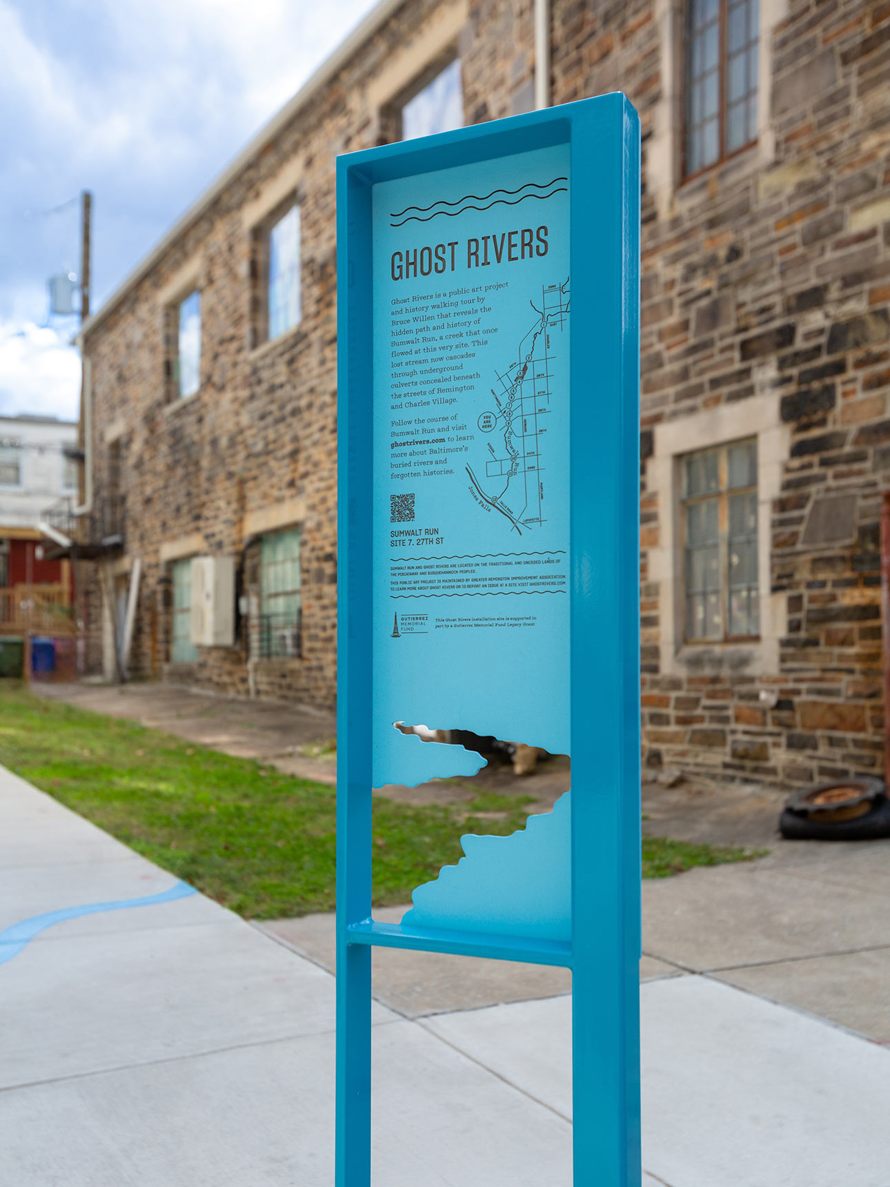 A photo of a tall blue interpretive sign with the form of a river cut out from the bottom of the sign panel. A large stone building is visible in the background.