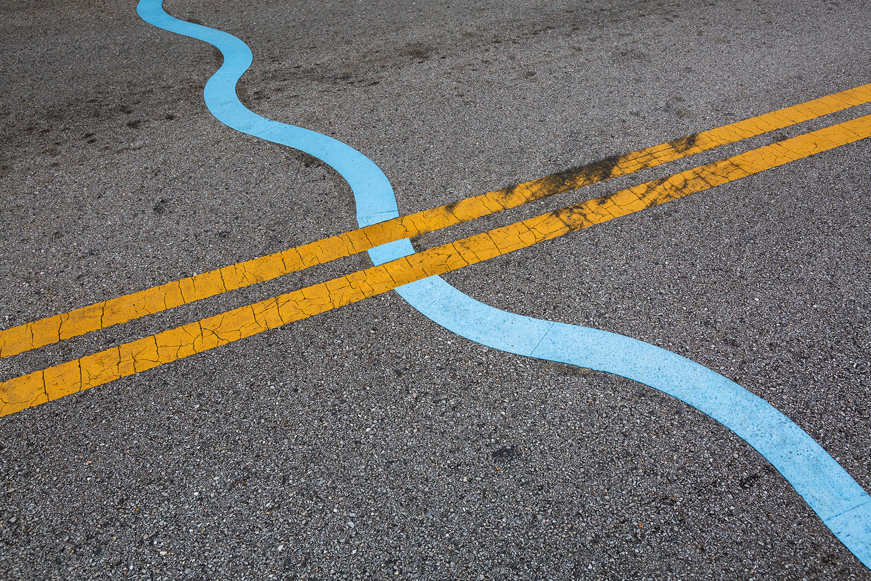 Detail photo of a bright blue six-inch wide wavy line on the street, intersecting with yellow road-marking lines.