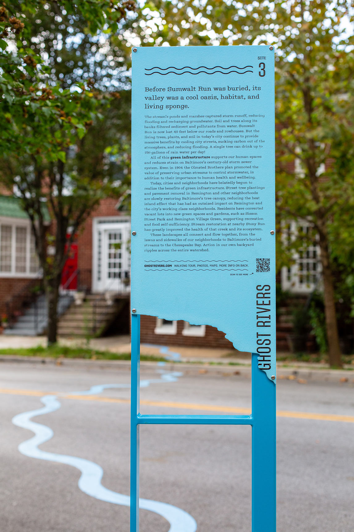 A photo of a tall blue interpretive sign, with the form of a river cut out from the bottom of the sign panel. Through the gap in the sign, a bright blue wavy line snakes across the street, with Baltimore rowhouses visible in the background.