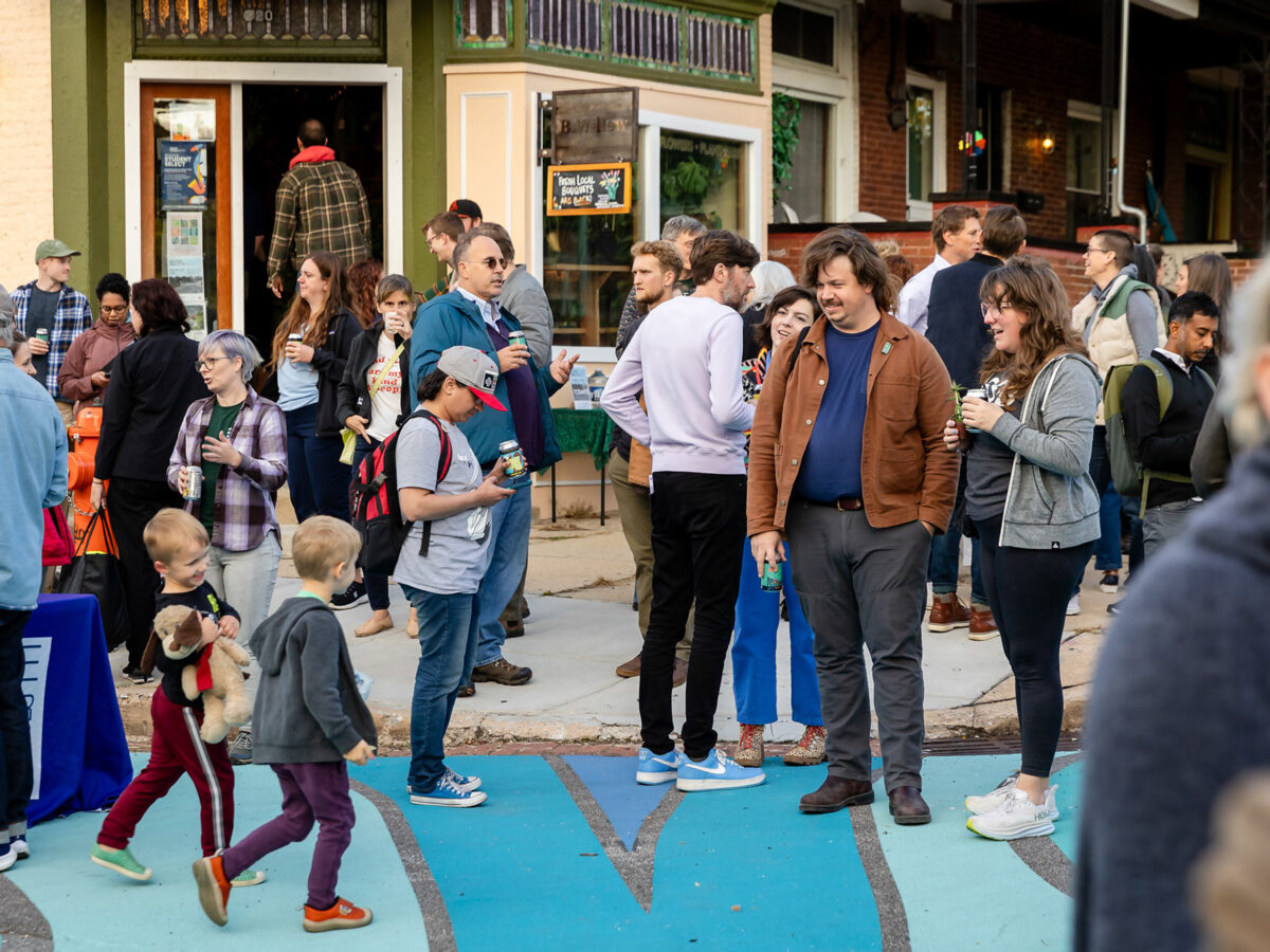 A crowd of enthusiastic neighbors and playing children gathers in front of a local plant shop at the opening ribbon-cutting celebration for Ghost Rivers public art installation.