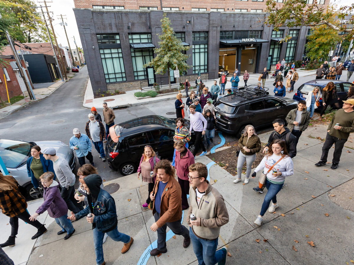 A large crowd of people crosses the street, following the trail of Sumwalt Run and the Underground Water Goddess at the opening ribbon-cutting celebration for Ghost Rivers public art installation.