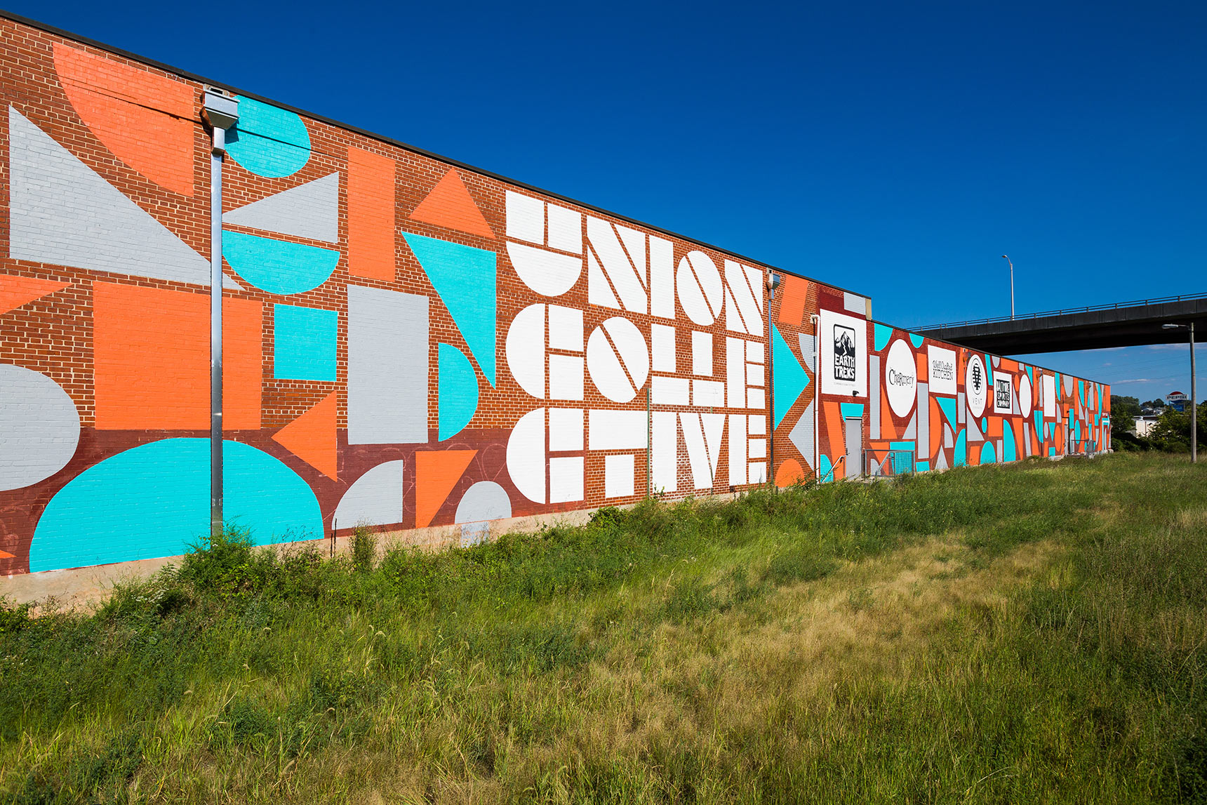Enormous mural on the side of the Union Collective warehouse building in baltimore composed of colorful orange, blue, and grey geometric shapes including circles, triangles, parallelograms, squares, etc.