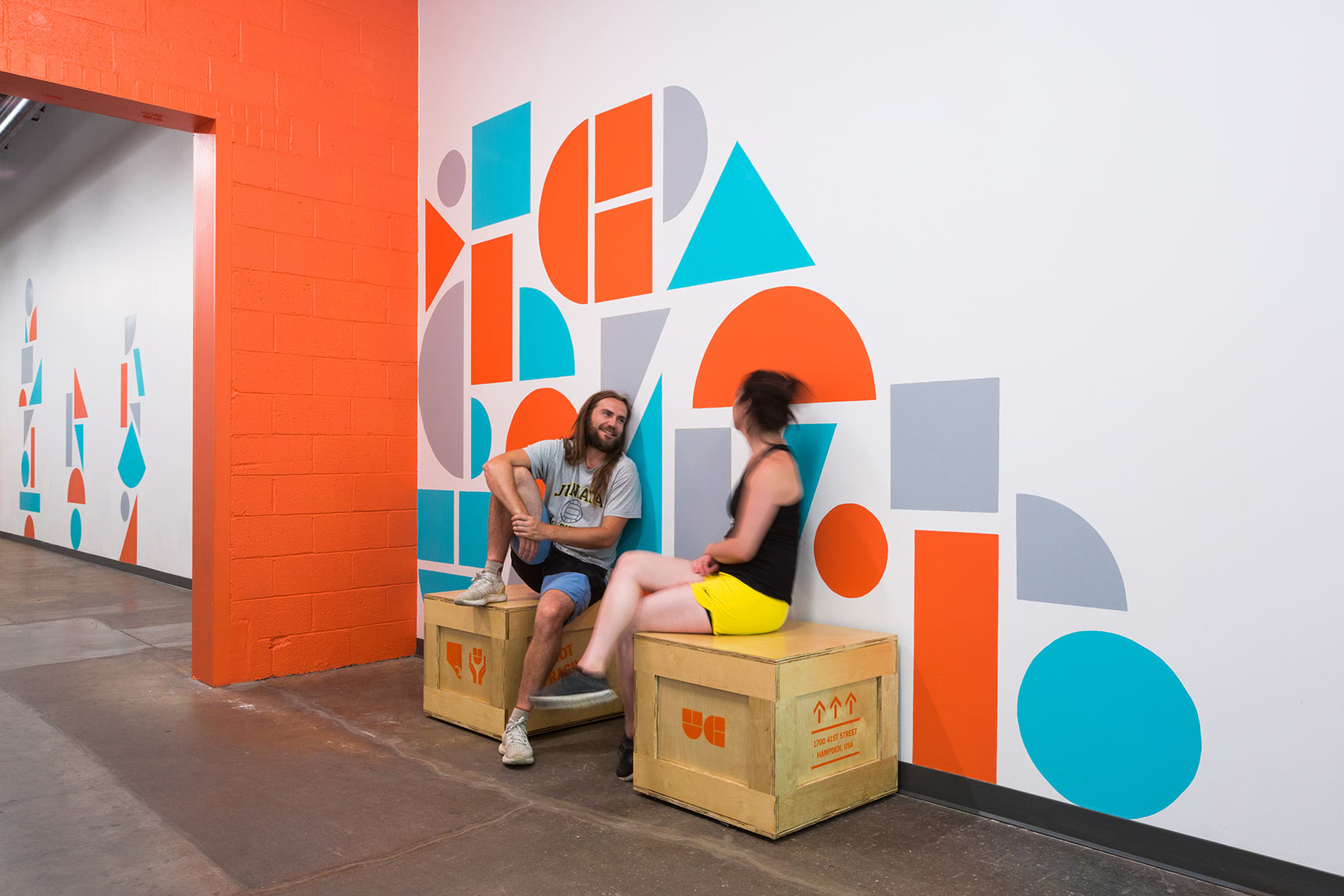 People sitting on custom shipping crate seating in front of colorful geometric shape supergraphic murals in the hallways of the Union Collective building